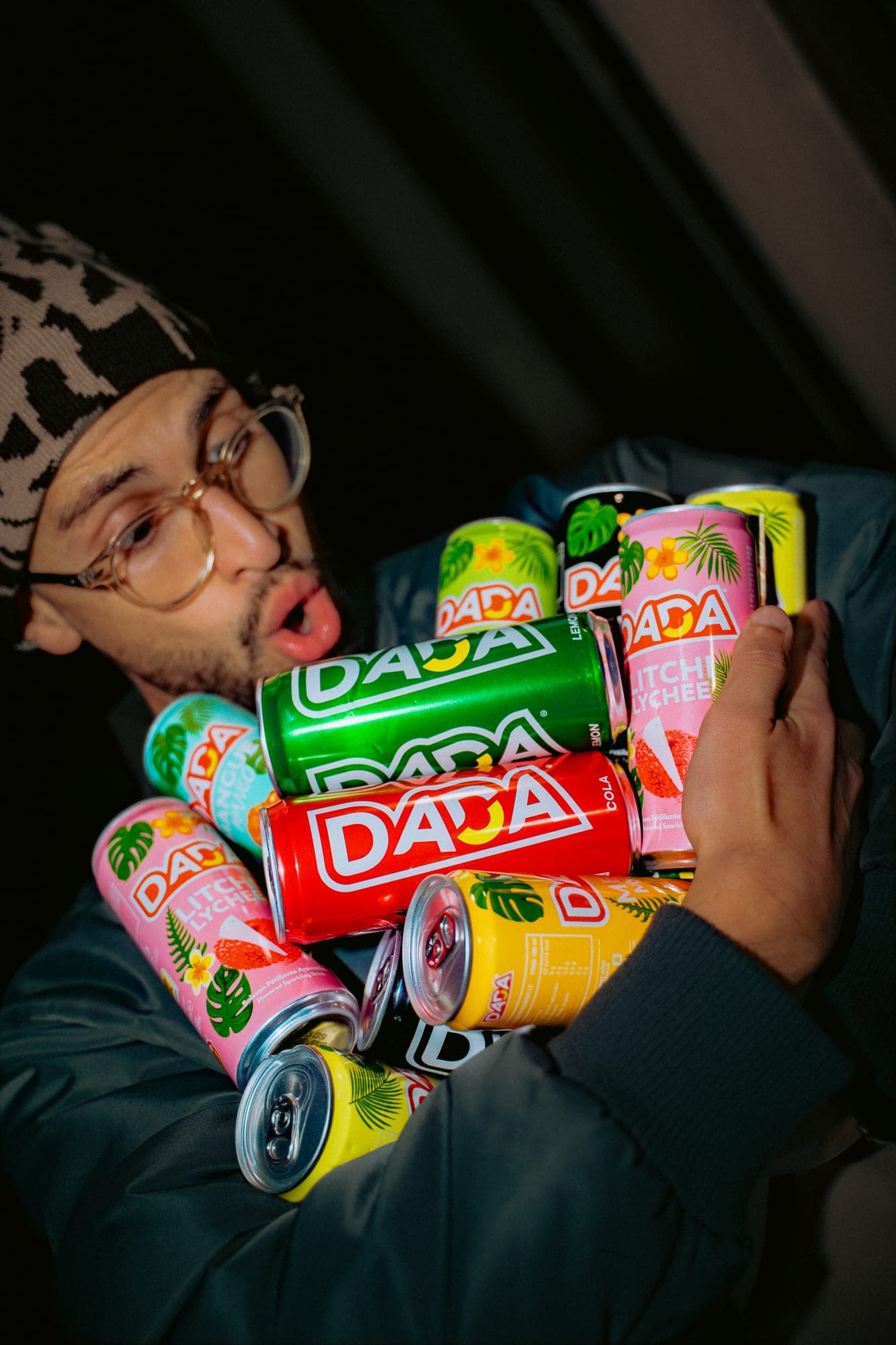 Homme au bonnet portant une brassée de canettes Dada colorées, dans une lumière basse