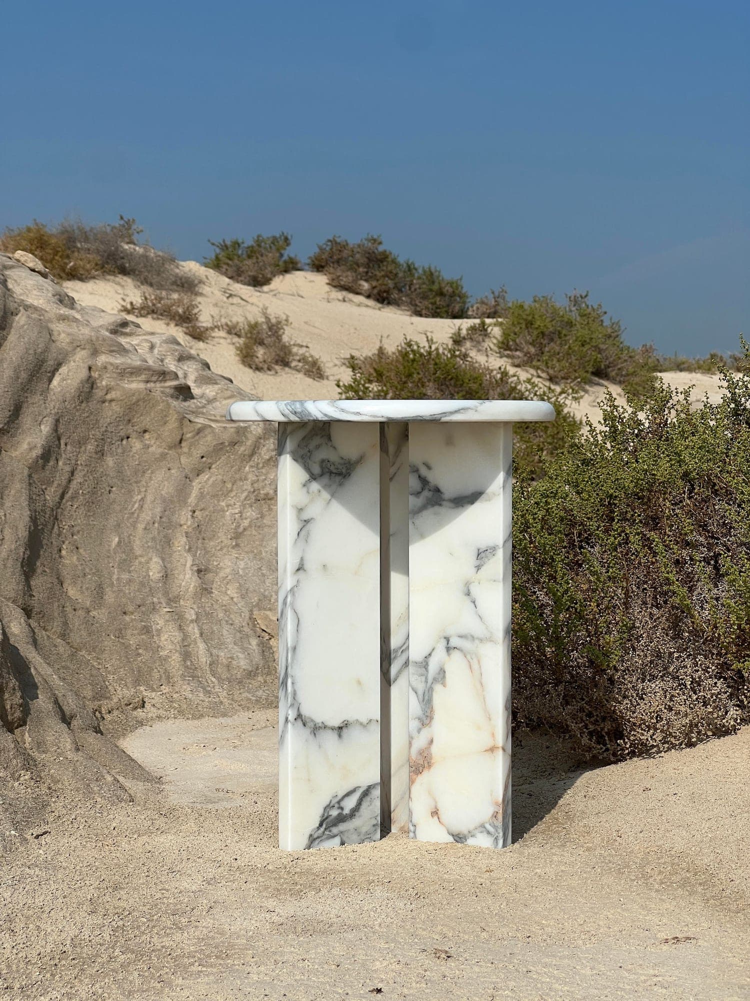 Table basse en marbre blanc veiné posée sur le sable, devant un rocher et des buissons