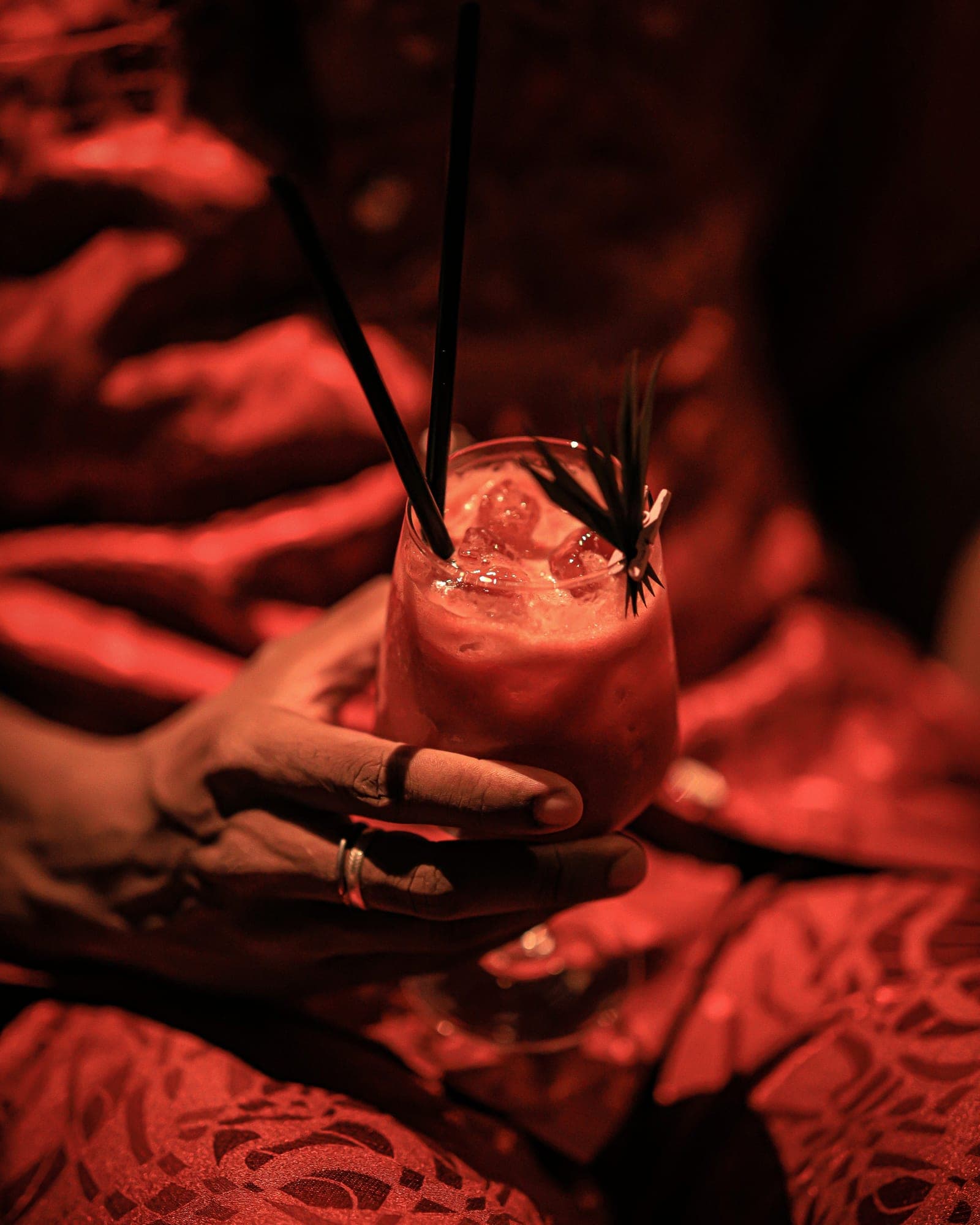 Mains baguées tenant un cocktail glacé sur une nappe brodée, dans une lumière rouge